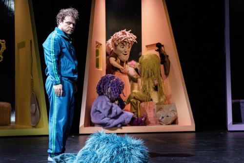 Bühnensituation: Parsa Yaghoubi Pour in einem blauen Trainingsanzug schaut auf ein flauschiges blaues Stoffstück auf dem Boden. Im Hintergrund ein kleiner, dreieckiger hoher orangefarbener Mini-Raum, in dem sich drei Personen mit großen Kopfmasken befinden.