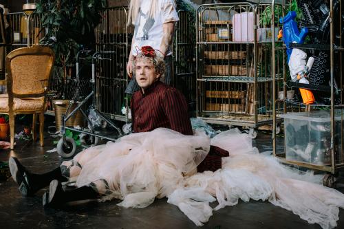 Christoph Jöde sitzt auf dem Boden. Sein Mund ist geöffnet. Er trägt einen Rock und eine Schärpe aus Tüll, schwarze Kniestrümpfe und schwarze Schuhe. Sein Jackett mit Puffärmeln ist schwarz-rot gestreifte. Auf seinen blonden Locken trägt er einen Plastikhummer.  Hinter ihm steht Henni Jörissen, ihren Kopf sieht man nicht. Dahinter stehen vergitterte Rollwägen mit Requisiten, darunter Holzkisten, Blumenkübel, Pistolen und Plastikgewehre. Links im Bild steht ein Rollator und ein goldener Sessel im Barockstil.