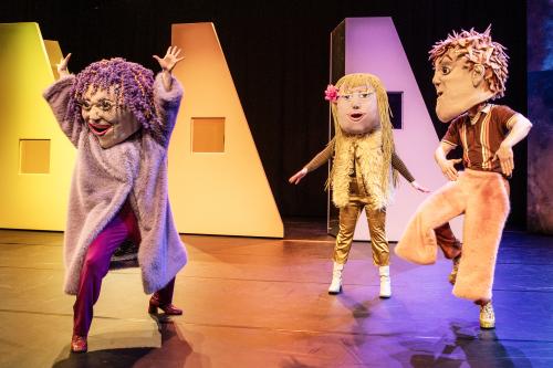 Bühnensituation: Drei Personen mit großen Kopfmasken nebeneinander. Sie tragen goldene, lolafarbene und rosarfarbene Kleidung, dazu passend ihre Haare in lila, gold und rosa. Sie haben die Arme seitlich oder nach oben ausgestreckt erhoben, stehen im Ausfallschritt oder haben ein Bein angehoben.