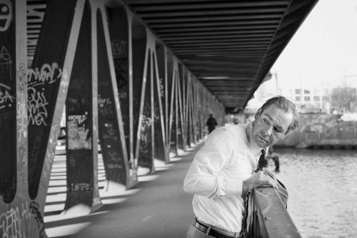 Außenansicht. Schwarz-Weiß-Bild. Mirco Kreibich steht leicht über ein Geländer gebeugt auf einer Brücke über einem Fluss. Links von ihm sind die Brückenaufbauten im Bild. Mirco dreht sich leicht über seine rechte Schulter und schaut hinter sich. Er trägt ein weißes Hemd, eine Krawatte, eine helle Hose mit Gürtel und einen großen Ring am rechten Zeigefinger. Seine Jacke hat er über das Geländer gelegt. Rechts im Bild ist das Wasser des Flusses im Bild.