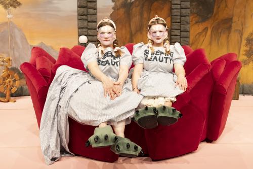 Bühnensituation: Christine Ochsenhofer und Anouk Piwek, beide in einem hellen gepunkteten und Kleid mit Puffärmeln sitzen auf einem roten Plüschsofa mit Zacken, die Hände in den Schoß gelegt. Auf ihrer Brust steht „SLEEPING BEAUTY“ in schwarzen Großbuchstaben. Ihre langen blonden Haare sind eingedreht und mit rosafarbenen Bändern festgebunden. Sie tragen Face-Lift-Masken, die Gesicht bis auf Mund und Augen vollständig bedecken. An ihren Füßen befinden sich große grüne Puschen mit schwarzen Krallen daran.
