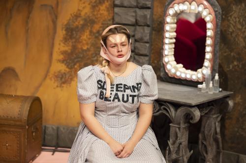 Bühnensituation: Anouk Piwek in einem hellen gepunkteten Kleid mit Puffärmeln, sitzt. Auf ihrer Brust steht „SLEEPING BEAUTY“ in schwarzen Großbuchstaben. Ihre langen blonden Haare sind eingedreht und mit rosafarbenen Bändern festgebunden. Sie trägt eine Face-Lift-Maske, auf ihre Stirn sind drei helle Pflaster geklebt. Im Hintergrund steht ein Schminktisch in Stein-Optik, um dessen Spiegel sich eine riesige Zahnreihe mit einer leuchtenden Zahnspange befindet.