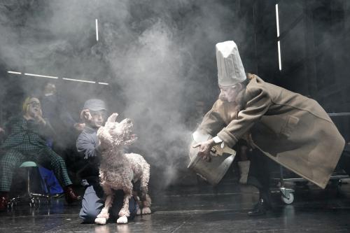 Im Vordergrund ist Felix Knopp zu sehen, er trägt eine weiße Kochmütze und einen langen beigen Mantel und hält einen großen Kochtopf, aus dem es stark qualmt. Links von ihm ist eine lebensgroße Hundepuppe zu sehen, die von Oscar Olivo gespielt wird. Letzterer hält den Hundekopf und ist in blauem Overall und farblich passender Schirmmütze gekleidet. Im linken Hintergrund sitzt Bettina Stucky in grün-schwarz kariertem Hosenanzug und gelber Perücke auf einem Drehstuhl.  