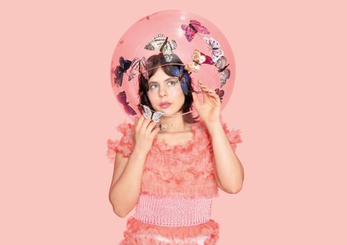 Eine weiblich gelesene Person in rosa Rüschenkleid vor rosa Hintergrund ist zu sehen. Um ihren Kopf ist eine durchsichtige Blase, in der Schmetterlinge schweben.