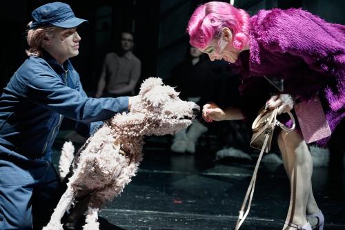 Oscar Olivo im blauen Overall und blauer Schirmmütze und hellen Haaren darunter hat seine Hand an einer lebensgroßen Hundepuppe mit hellem Fell. Der Hund hat seine Schnauze an der Hand von Angelika Richter, die rechts im Bild zu sehen ist und sich zum Hund herunterbeugt. Sie trägt eine pinke Perücke mit Locken, dazu einen lila Fellmantel und eine bronzefarbene Handtasche. Sie lächelt breit. 