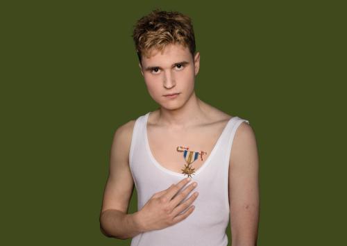 Eine männlich gelesene Person mit blonden kurzen Locken in weißem Tanktop ist zu sehen. Eine Hand deutet auf eine Brosche, die an ein Abzeichen erinnert und in die Haut sticht. Die Person schaut in die Kamera. Der Hintergrund ist dunkelgrün.