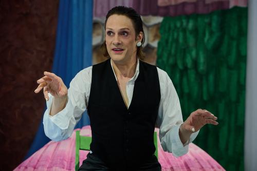 Less Brandes-Hell trägt ein weißes Hemd, darüber eine schwarze Weste, ihre Fingernägel sind weiß lackiert. Sie trägt pinken Lippenstift, große weiße Ohrringe und ihre Augen sind in zartem lila geschminkt und sind weit aufgerissen. Die Arme sind etwas nach vorgestreckt, sie gestikuliert, ihr Mund ist leicht geöffnet. Im Hintergrund sieht man verschiedene bunte Stoffe in pink, blau, grün, lila. 