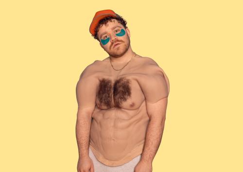 Eine männlich gelesene Person mit gelber Cap und braunen Locken und blauen Augenpads schaut in die Kamera. Über seinem Oberkörper hängt ein menschlicher Oberkörper mit behaarter Brust und Sixpack aus Silikon. Er trägt eine goldene Kette, der Hintergrund ist gelb. 