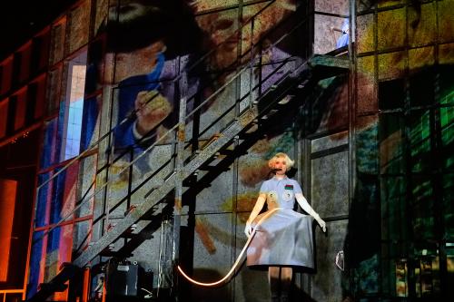 Sandra Gerling ist in der Mitte der Bühne zu sehen, sie hat kurze blonde Locken, trägt ein graues Kleid mit einem runden Reifrock und spitzen Kappen vor den Brüsten und hat einen schwarzen Schlauch im Mund. Ein Kabel ragt auf der Höhe ihres Bauchs in ihr Kleid. Der Rest der Bühne ist dunkel. Man kann eine schwarze Treppe oberhalb von Sandra Gerling erkennen und eine Art Wand aus Kacheln. An die Wand wird ein Video projiziert, auf dem schemenhaft Gesichter zu erkennen sind.