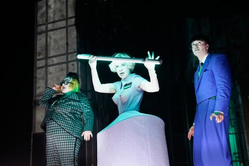 Vor einem schwarzen Hintergrund stehen drei Personen, alle sehen nach vorn. Bettina Stucky trägt einen grün-schwarz karierten Hosenanzug, eine blonde Perücke und eine große Brille mit schwarzem Rahmen. Sandra Gerling steht mittig, hat einen kurzen blonden Bob, trägt ein graues Kleid mit einem Reifrock und spitzen Kappen vor den Brüsten. In den Händen hält sie eine Neonröhre. Rechts steht Maximilian David Scheidt in blauem Anzug, seine Haare sind schwarz. Alle Münder sind leicht geöffnet.