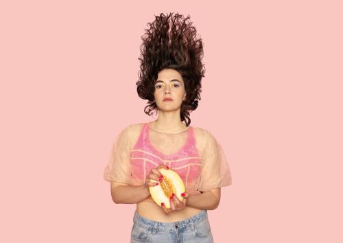 Eine weiblich gelesene Person hat braune Locken in die Luft geworfen. Sie trägt einen pinker Sport-BH, darüber ein durchsichtiges T-Shirt und eine Jeans. Ihre pinken Fingernägel greifen in eine aufgeschnittene Melone. Der Hintergrund ist pink.