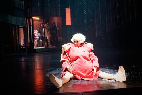 Sandra Gerling sitzt mit ausgestreckten Beinen auf dem Bühnenboden und schaut nach links. Sie trägt eine blonde Bob-Perücke, ihre Augen sind schwarz geschminkt. Sie trägt eine übergroße pinke Uniform mit goldenen, fransigen Epauletten auf den Schultern und zwei Knopfreihen, ebenfalls in Gold. Ihre Beine sind nicht bedeckt, sie trägt weiße Sneaker. Im dunklen Hintergrund sieht man verschwommen eine Gruppe Menschen in einem kleinen, schwach erleuchteten Raum stehen. 