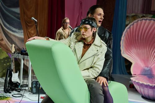 Bühnensituation: Juno Peter und Less Brandes-Hell sitzen hintereinander auf einem grünen Sofa, dessen lange Seite in die Luft gekippt ist. Beide tragen Lederjacken mit Fransen, Juno sitzt vorn und trägt ein Bandana-Tuch um die Haare und eine Fliegersonnenbrille. Junos Lippen sind gespitzt, their rechte Hand liegt auf der Sofaspitze. Im Hintergrund steht Micha Krause an einem Mikrofon, eine E-Gitarre ist zu sehen und eine große aufgeklappte rosa Muschel.