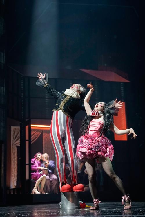 Im Vordergrund sind Felix Knopp und Sachiko Hara zu sehen. Er trägt eine grau-weiß-rote enge Hose und rote Clownsschuhe, eine schwarze Weste mit Pailletten und eine weiße Krause um den Hals. Augen und Mund sind weit aufgerissen, er schaut nach oben und hat beide Arme seitlich ausgestreckt. Sachiko Hara trägt ein Kleid, das mit Rosen besetzt ist. Ihre Krause ist rosa, ihre langen schwarzen Haare sind offen. Sie schaut hoch, Mund und Augen weit auf und die Arme sind nach oben und zur Seite gestreckt. 