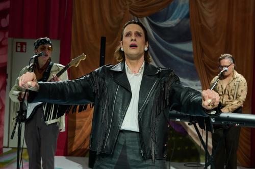 Bühnensituation: Less Brandes-Hell trägt eine schwarze Fransen-Lederjacke, ihre Arme sind nach vorne ausgestreckt, als würden sie etwas umfassen. Ihr Mund ist geöffnet, sie schaut nach vorn. Im Hintergrund stehen Juno Peter mit E-Gitarre und Micha Krause am Keyboard, Juno singt in ein Mikrofon und trägt ebenfalls eine Lederjacke.