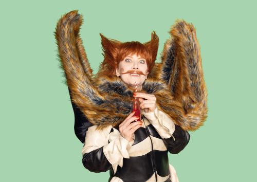 Zu sehen ist eine Person mit orangenen Haaren, die zu zwei Hörnern oder Ohren nach oben frisiert sind. Die Augen sind weit aufgerissen, sie trägt eine orangene Flasche mit einem gestreiften Strohhalm darin in den Händen. Außerdem trägt die Person einen orangenen Schnurrbart und zwei Fangzähne blitzen hervor Eine Art Fellponcho ist zu beiden Seiten nach oben geklappt. Darunter trägt die Person einen schwarz-weißen, ledrigen Anzug. Der Hintergrund ist grün.