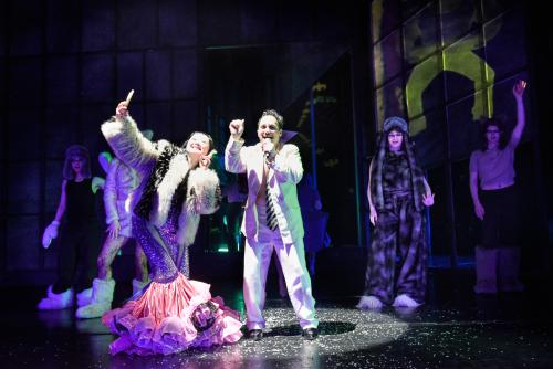 Im Vordergrund stehen Sachiko Hara und Oscar Olivo. Sie trägt eine Felljacke und darunter einen Rock aus lila Pailletten mit Tüll ab den Knien, sodass die Füße verdeckt sind. Sie hält Zeige- und Mittelfinger hoch und macht ein Selfie mit einem Handy. Sie lächelt. Oscar Olivo trägt einen weißen Anzug und eine schwarz-gelb gestreifte Krawatte. Er hat eine Hand in die Höhe gestreckt, mit der anderen hält er ein Mikrofon an seinen geöffneten Mund. Im dunklen hinteren Bühnenbereich sieht man die Chormitglieder. 