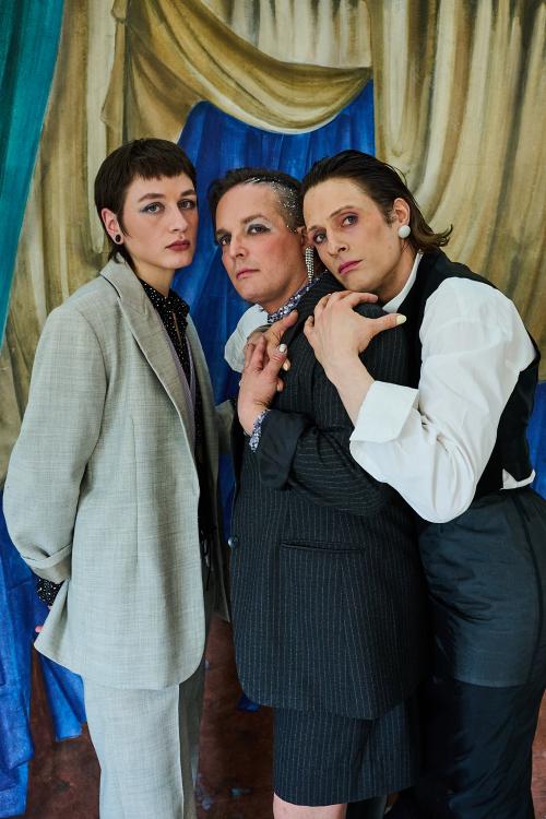 Juno Peter, Micha Krause und Less Brandes-Hell stehen eng nebeneinander und schauen in die Kamera. Less hat ihren Kopf und die Arme um Michas Schultern gelegt, Micha berührt ihre Hand. Sie stehen vor einem gemalten gelben Vorhang.