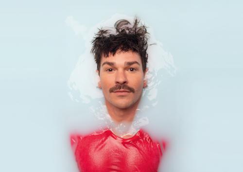 Eine Person mit braunen Haaren und Schnurrbart schaut gerade in die Kamera. Sie scheint in einer blauen Flüssigkeit zu versinken oder aus ihr aufzutauchen. Außerdem trägt sie ein pinkes Oberteil.