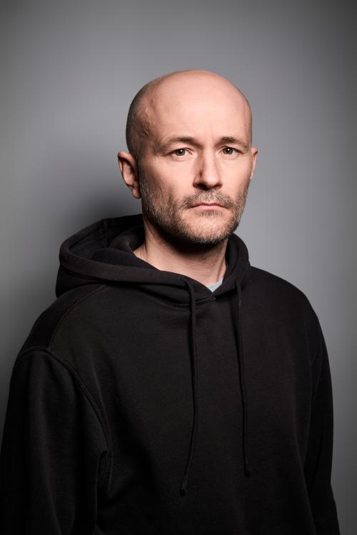 Es ist ein Porträtbild von Moritz Grove vor einem grauen Hintergrund. Er trägt ein schwarzes Oberteil.