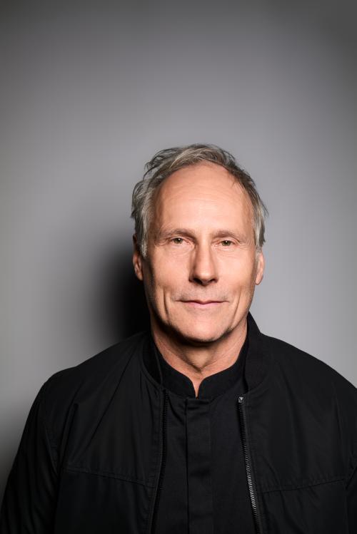 Es ist ein Porträtbild von Wolfram Koch vor einem grauen Hintergrund. Er trägt ein schwarzes Oberteil.