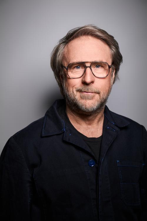 Es ist ein Porträtbild von Bjarne Mädel vor einem grauen Hintergrund. Er trägt ein schwarzes Oberteil.