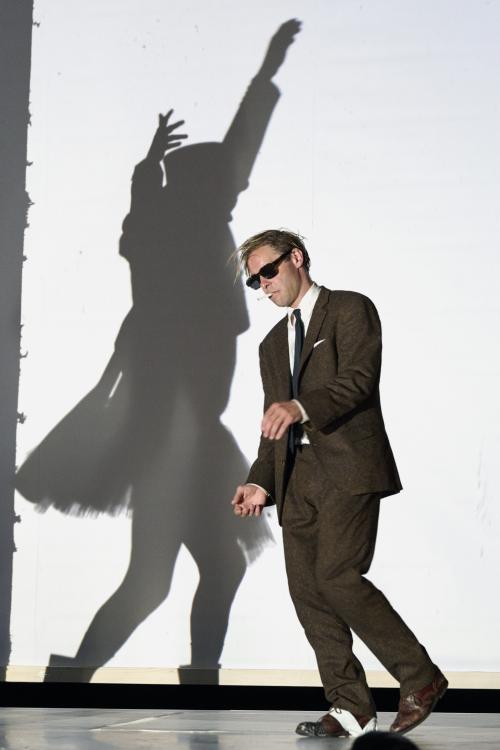 Bühnensituation: Mirco Kreibich mit braunem Anzug mit grüner Krawatte, Sonnenbrille und Zigarette im Mund, seine Arme und ein Bein sind leicht angewinkelt. Im Hintergrund der Schatten einer Person im kniefreien Kleid, die beide Arme erhoben hat, auf einer weißen Wand.