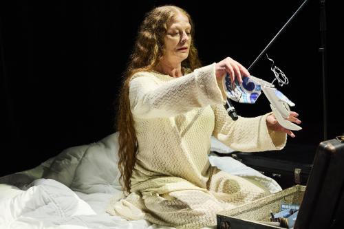 Christiane von Poelnitz sitzt auf einem weiß bezogenen Bett. Der Hintergrund ist dunkel. Sie trägt ein beiges Stickkleid. Vor ihr ist ein offener schwarzer Koffer. In den Händen hält sie eine blau weiße Bastelarbeit aus Keramik, die mit Draht verbunden ist.