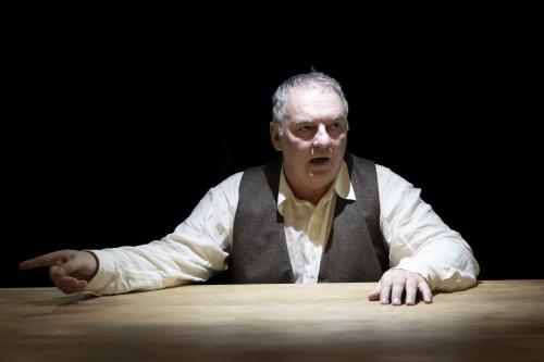 Hermann Book sitzt an einem Holztisch, nur sein Oberkörper und Kopf sind zu sehen. Unter einer braunen Weste trägt er ein helles Hemd. Eine Hand liegt auf dem Tisch, mit der anderen zeigt er nach links. Sein Mund ist geöffnet, er schaut nach rechts. Der Hintergrund ist schwarz. 