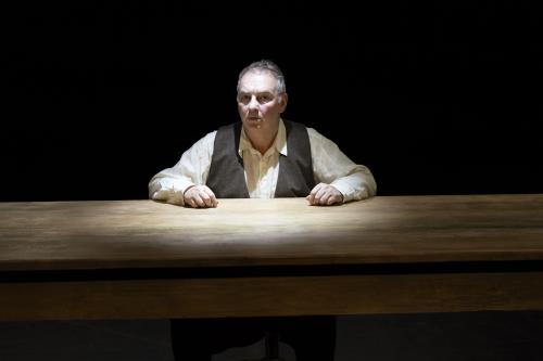 Hermann Book sitzt an einem Holztisch, nur sein Oberkörper und Kopf sind zu sehen. Unter einer braunen Weste trägt er ein helles Hemd. Beide Hände liegen auf dem Tisch. Sein Mund ist geschlossen, er schaut in die Kamera. Der Hintergrund ist schwarz. 