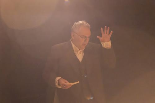 Hermann Book steht mit einem Blatt Papier in der Hand und der anderen offenen Hand erhoben vor einem schwarzen Hintergrund. Er hat den Mund geöffnet, schaut nach unten. Licht strahlt von oben und taucht die Szene in eine warme, braune Atmosphäre.  