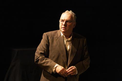 Hermann Book steht mit einem Blatt Papier in den Händen vor einem schwarzen Hintergrund. Er ist gekleidet in braunem Sakko und weißem Hemd darunter. Er hat den Mund leicht geöffnet, schaut nach links.  