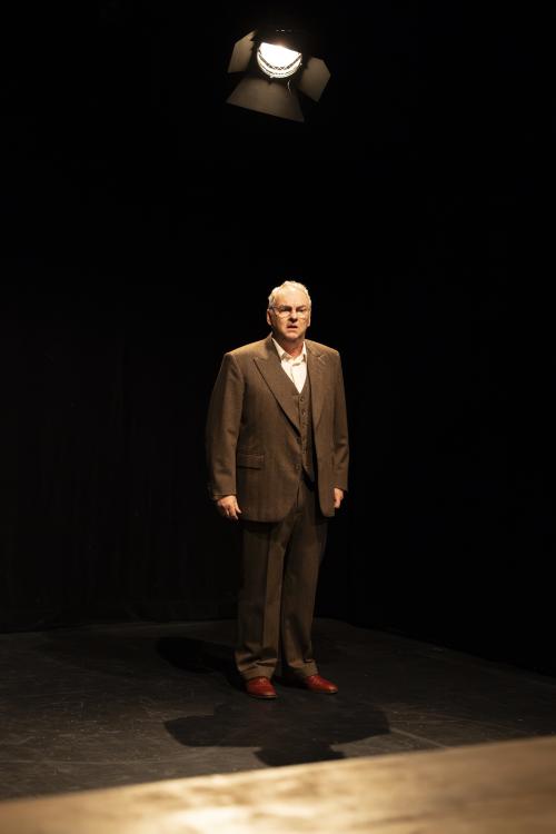 Hermann Book steht vor einem schwarzen Hintergrund auf schwarzem Boden. Gekleidet ist er in brauner Hose, braunen Lederschnürschuhen, brauner Weste und weißem Hemd darunter. Er trägt eine Brille, sein Mund ist leicht geöffnet, er schaut geradeaus. Über ihm hängt ein Scheinwerfer. 