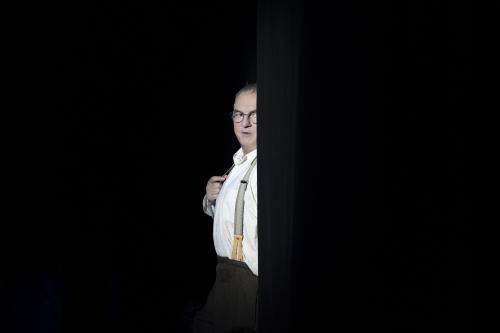 Hermann Book steht halb versteckt hinter einer schwarzen Wand, alles außer ihm ist dunkel. Er lugt hervor, trägt ein weißes Hemd, eine Brille und Hosenträger. 