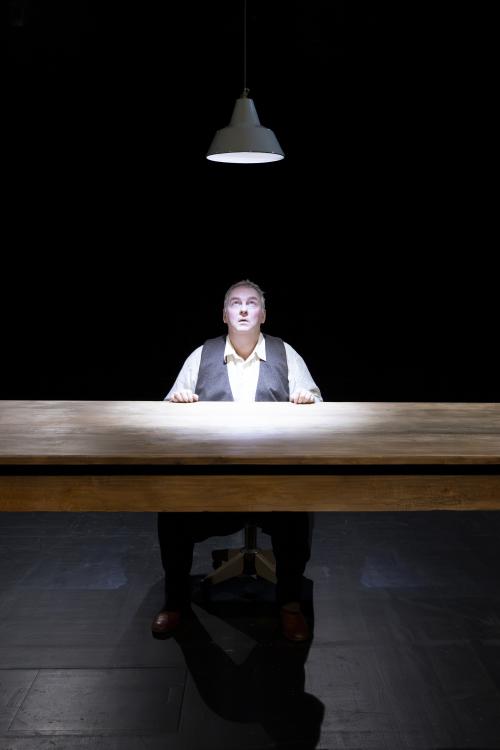 Hermann Book sitzt an einem Holztisch, über ihm leuchtet eine Hängelampe, zu der er aufblickt. Unter einer braunen Weste trägt er ein helles Hemd. Seine Hände liegen auf dem Tisch. Sein Mund ist geöffnet. Der Hintergrund und Boden sind schwarz. 