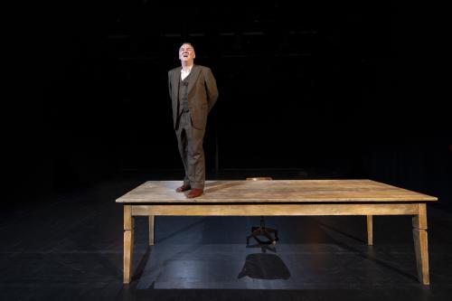 Ein großer rechteckiger Holztisch steht auf schwarzem Boden, vor einem schwarzen Hintergrund. Darauf steht Hermann Book, gekleidet in brauner Hose, braunen Lederschnürschuhen, brauner Weste und weißem Hemd darunter. Er schaut nach oben, sein Mund ist geöffnet. Hinter dem Tisch steht ein Holzstuhl.  