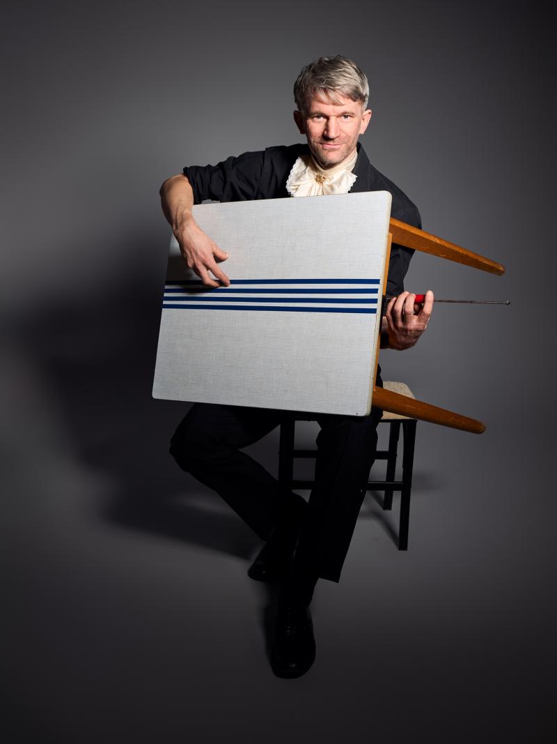 Der Schauspieler Friedrich Paravicini, er hält einen kleinen Esstisch wie eine Gitarre im Arm, Klebestreifen bilden die Saiten der Gitarre / fotografiert von Axel Martens