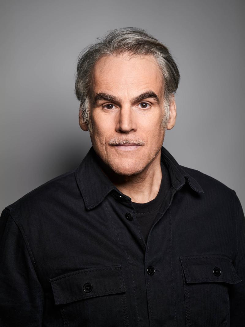 Es ist ein Porträtbild von Sebastian Blomberg vor einem grauen Hintergrund. Er trägt ein schwarzes Oberteil.