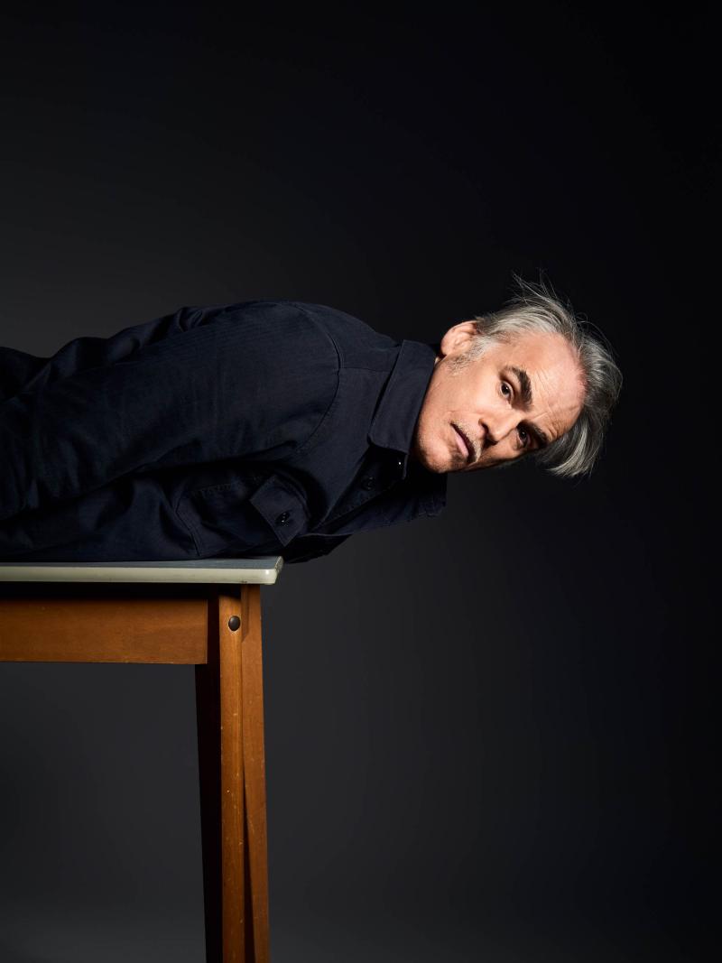 Sebastian Blomberg liegt auf einem Tisch. Es ist nur die Hälfte des Tisches mit Tischbein und Tischplatte zu sehen. Von Sebastian Blomberg ist nur der Oberkörper zu sehen. Brust und Kopf hängen über die Tischplatte hinaus. Er hat den Oberkörper und Kopf angehoben und schaut in die Kamera.