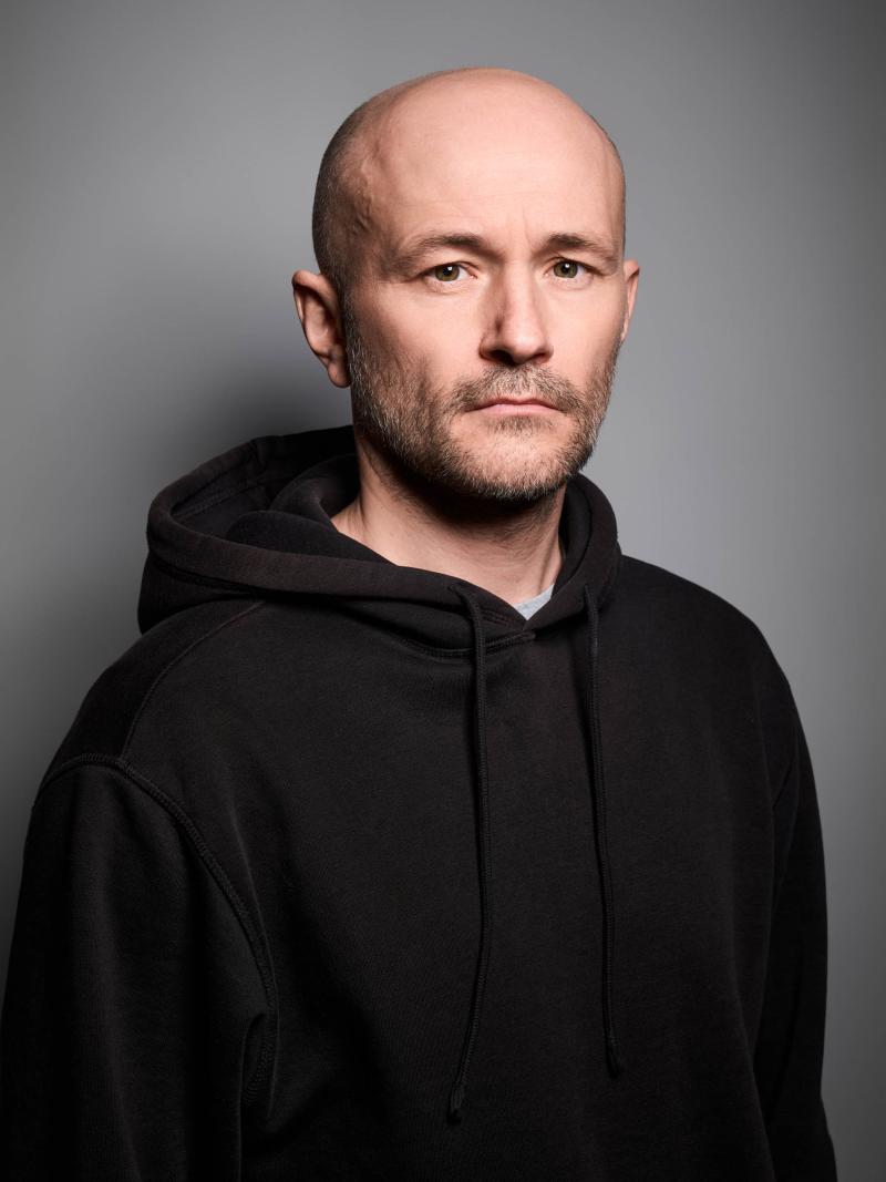 Es ist ein Porträtbild von Moritz Grove vor einem grauen Hintergrund. Er trägt ein schwarzes Oberteil.