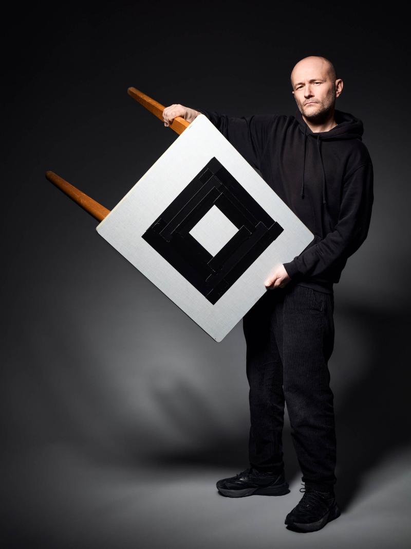 Ein Foto von Moritz Grove. Moritz Grove ist in der Totalen abgebildet. Er hält einen Tisch in beiden Händen und dreht die Tischplatte in Richtung der Kamera. Er schaut in die Kamera. Die Tischplatte ist mit weißem Stoff überzogen. Darüber bildet ein weiterer schwarzer Stoff eine Raute auf dem weißen Untergrund.