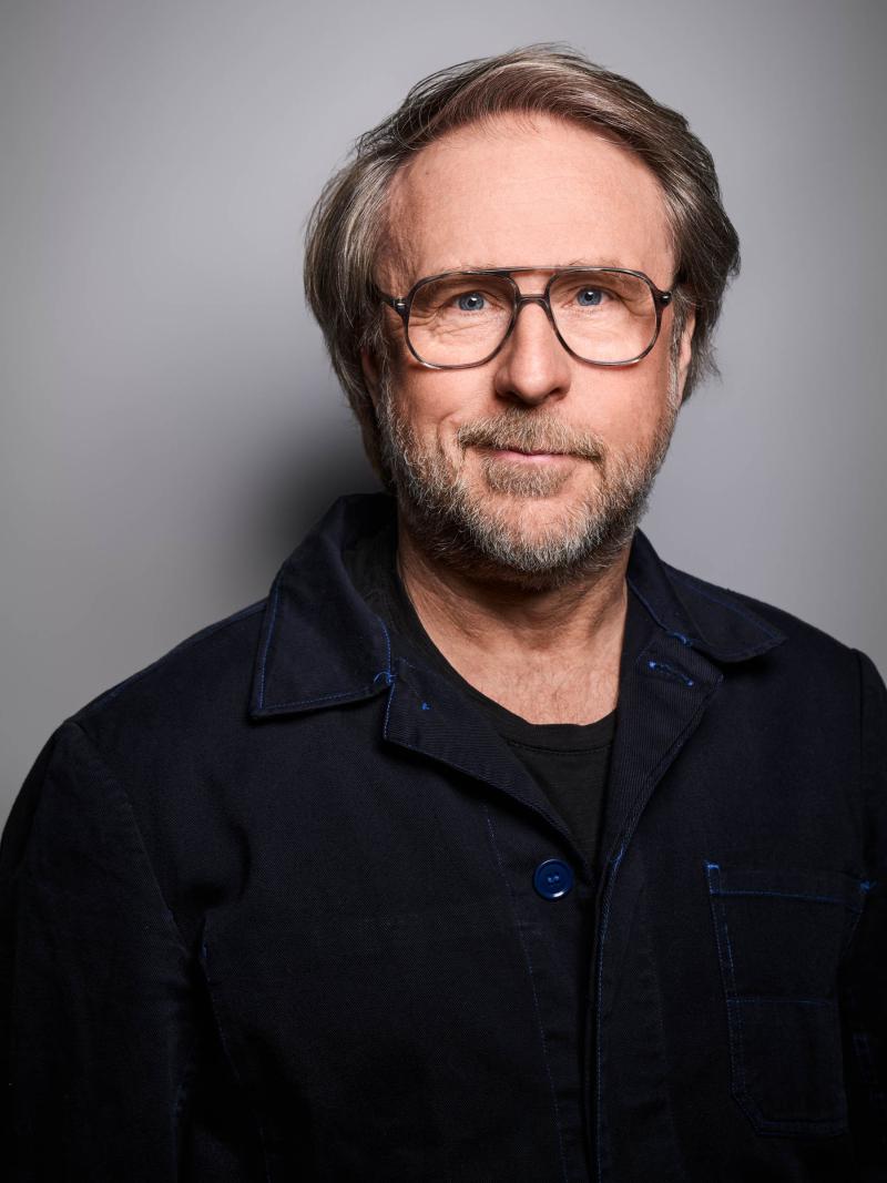 Es ist ein Porträtbild von Bjarne Mädel vor einem grauen Hintergrund. Er trägt ein schwarzes Oberteil.