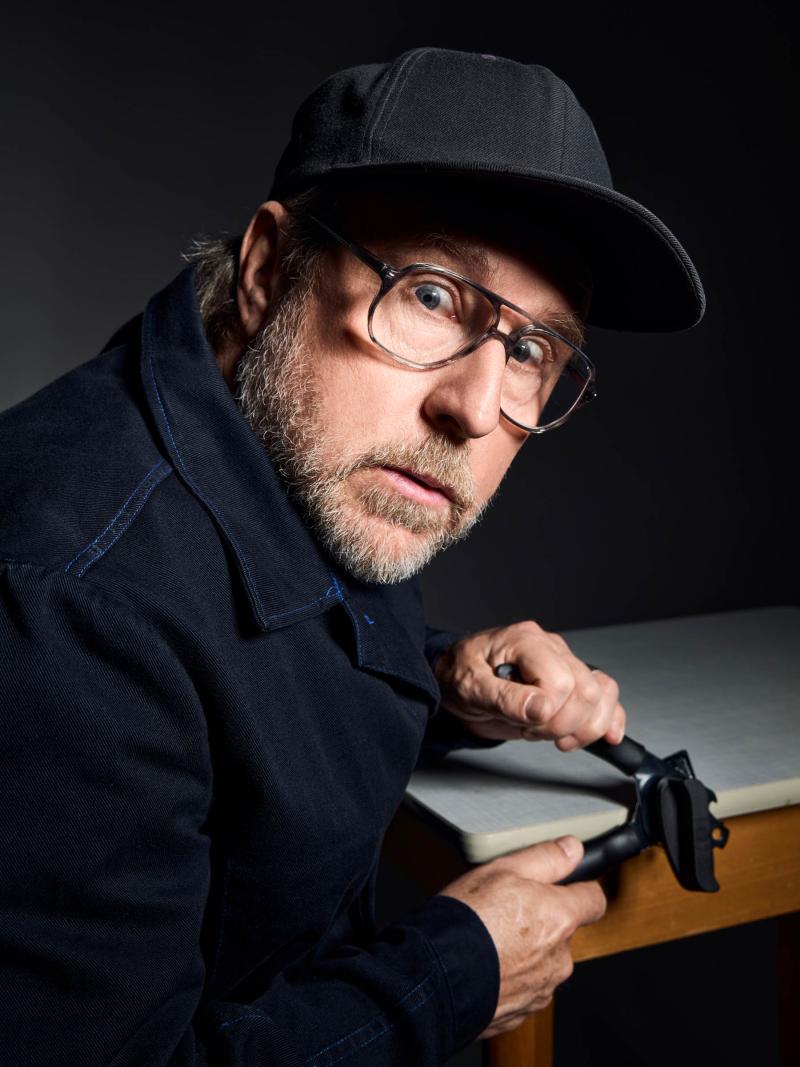 Es ist ein Foto von Bjarne Mädel. Er sitzt an einem Tisch und schaut über seine rechte Schulter erstaunt in die Kamera. Er trägt eine Brille und eine dunkle Cap. Er hält einen Dosenöffner an den Tisch und tut so, als würde er ihn damit öffnen.