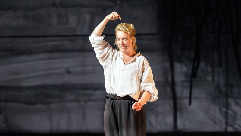 Lina Beckmann in weißer Leinenbluse und schwarzer Hose steht auf der Bühne, hebt den Arm über den Kopf und zeigt auf sich selbst. Ihr Ausdruck ist entschlossen und kämpferisch. Der Hintergrund zeigt eine dunkelgraue, abstrakt strukturierte Bühnenwand.