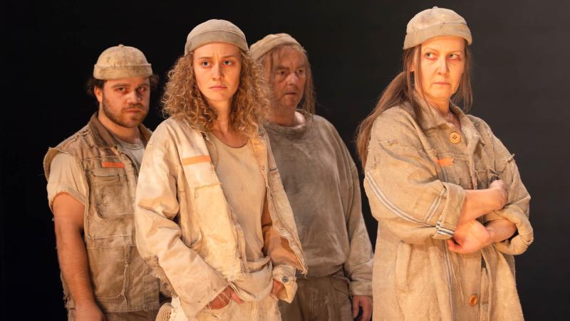 Vier Schauspieler*innen in zerschlissenen, erdfarben Kleidungsstücken und grauen Mützen stehen vor schwarzem Hintergrund und blicken ernst.