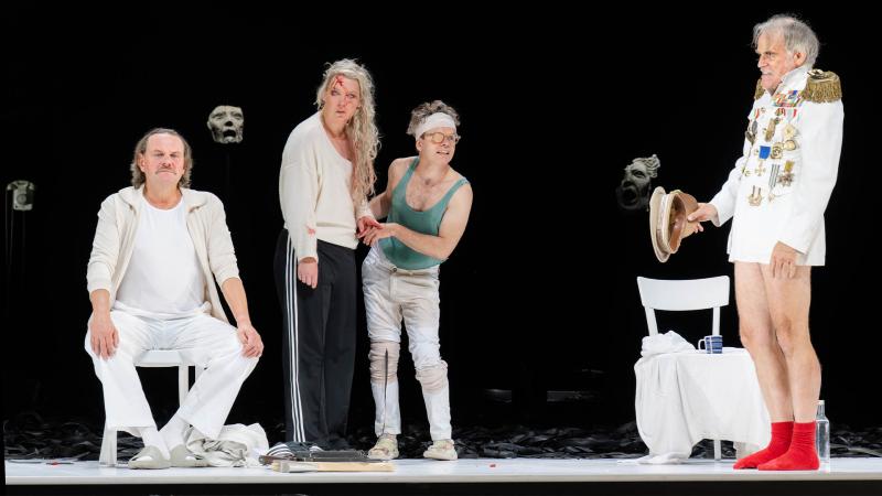 Devid Striesow, Lina Beckmann, Kristof van Boven und Sebastian Blomberg stehen auf einer weißen Bühne vor schwarzem Hintergrund mit schwebenden Masken. Links Devid Striesow in weißem Jogginganzug. Daneben Lina Beckmann und Kristof van Boven in Alltagskleidung – eine mit zerzaustem Haar und Blutspuren an der Stirn, der andere mit Stirnband und Augenklappe. Rechts steht Sebastian Blomberg in einem pompösen Militäruniform-Oberteil, roten Socken und ohne Hose, der einen Hut in der Hand hält.