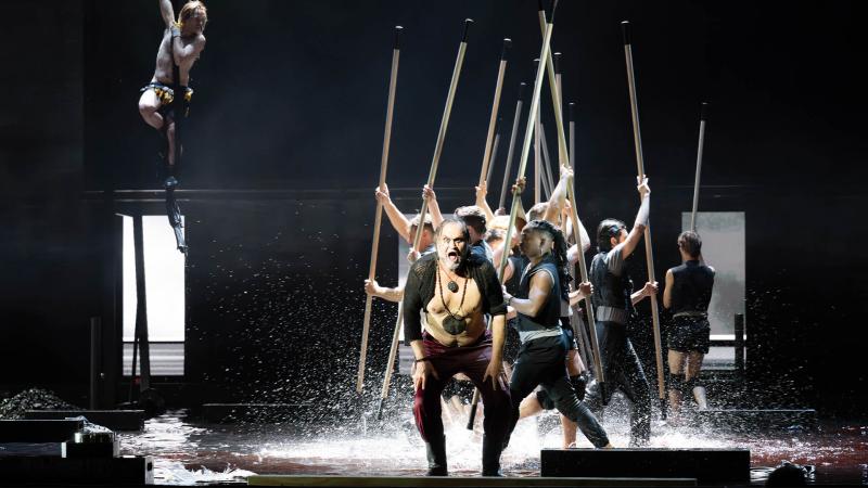 Theaterszene auf einer Bühne mit Wasser: Im Vordergrund Markus John mit freiem Oberkörper, roter Hose und Kettenanhänger, der mit weit geöffnetem Mund ruft oder singt. Hinter ihm eine Gruppe in Schwarz gekleideter Performer, die lange Stäbe schräg in die Höhe recken. Links oben hält sich Kristof van Boven oberkörperfrei in einem Vertikaltuch.