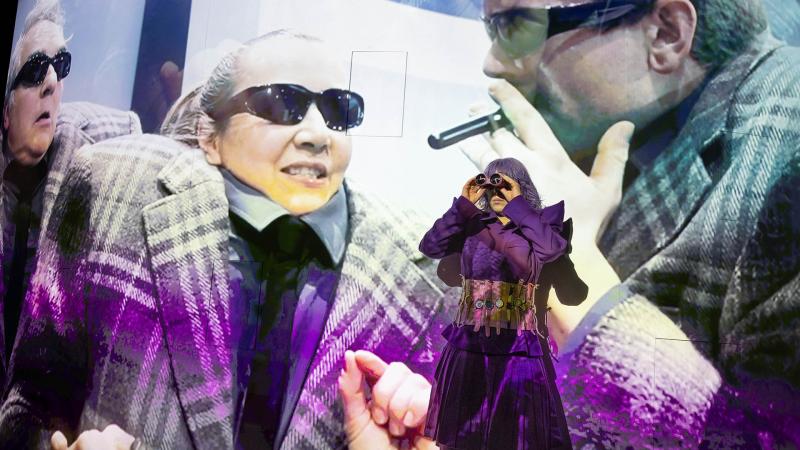 Eine Schauspielerin in violettem Rüschenkleid und Korsett blickt durch ein Fernglas, während hinter ihr eine großformatige Videoprojektion andere Schauspieler*innen mit Sonnenbrillen zeigt.