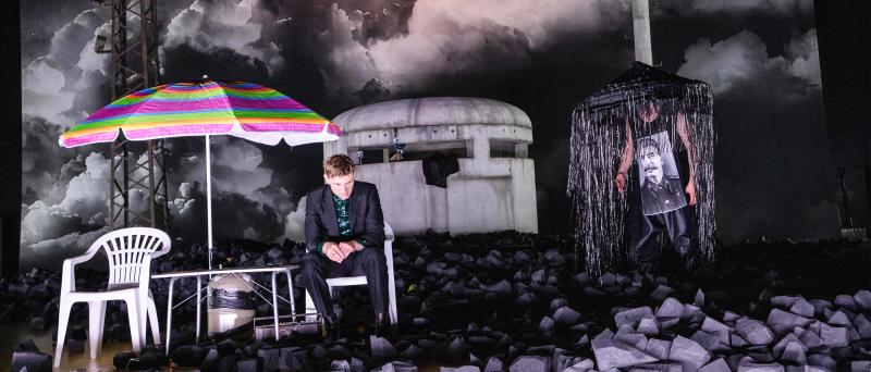 Der Schauspieler Paul Behren sitzt auf einem weißen Plastikstuhl unter einem bunten Sonnenschirm. Um ihn überall schwarze Kohle um ihn herum. Im Hintergrund dunkle Wolken am Himmel und ein gigantisches Schild, das verkehrt herum steht und von der anderen Seite den Schriftzug "Europe" zeigt.
