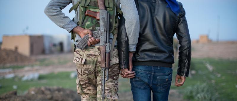 Ankündigungsmotiv "Über Grenzen gehen": Zwei Männer von hinten, sie halten sich an den Händen, einer trägt Tanrkleidung und eine Waffe auf dem Rücken